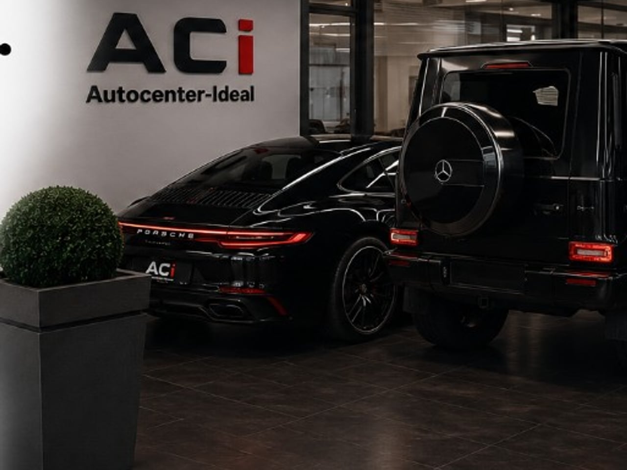 Showroom von Autocenter IDEAL mit Porsche 911 und Mercedes G-Klasse vor der ACI-Logowand